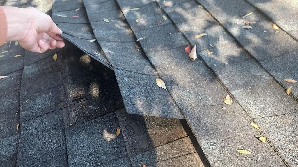 A hand lifts a loose asphalt shingle on a sloped roof, showing the gap underneath