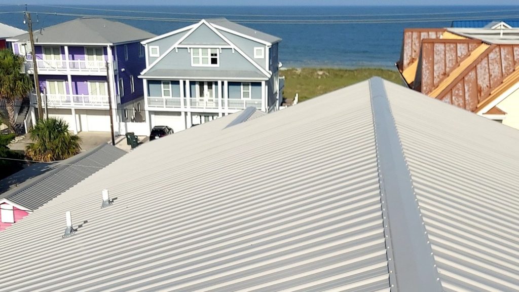 Light gray ribbed metal roof panels meet at a ridge cap, with beach houses and the ocean in the background