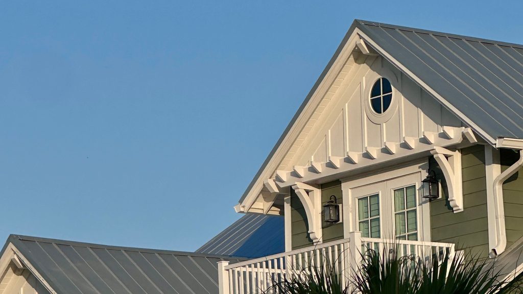 Coastal-style house with standing-seam metal roofs, white gable trim, a round attic window and a palm in front, under a clear blue sky