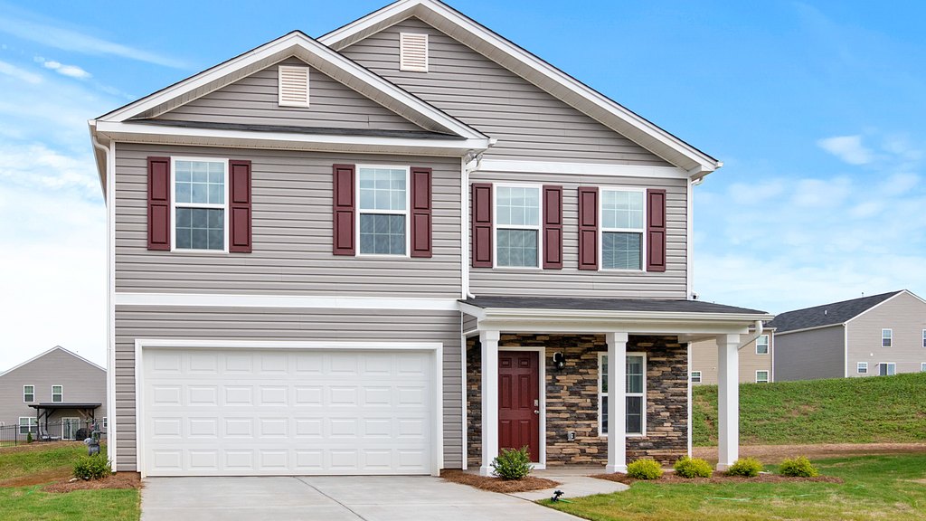 Two-story suburban home with gray vinyl siding, red shutters, a stone-front porch, an attached two-car garage and a dark shingle roof under a blue sky