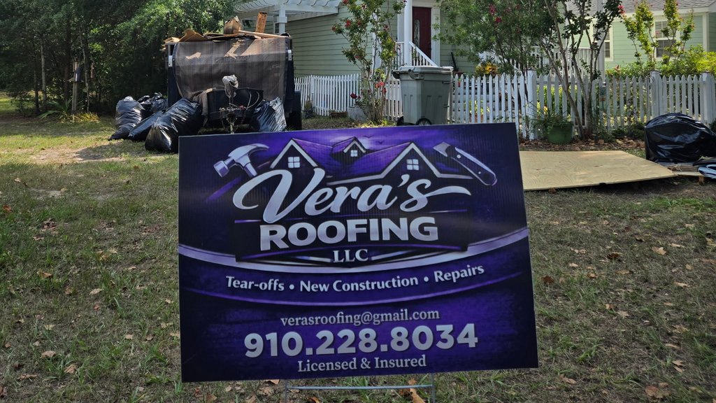 Purple Vera's Roofing yard sign on a lawn, with a loaded debris trailer, bagged roofing debris and a two-story green house behind it