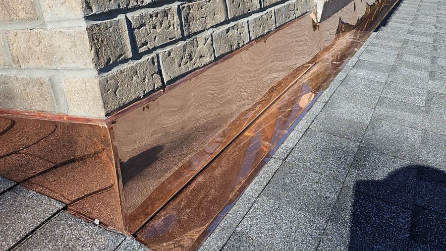 Copper flashing runs along the base of a brick wall where it meets a gray shingle roof