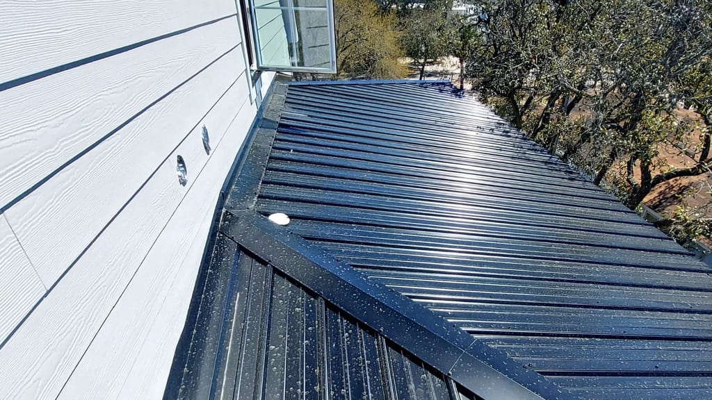 Dark ribbed metal roof panels run along a white lap-siding wall, with a metal trim piece where two panel runs meet